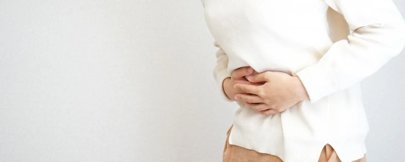 Une personne portant un pull blanc se tient le ventre, montrant un malaise sur un fond uni