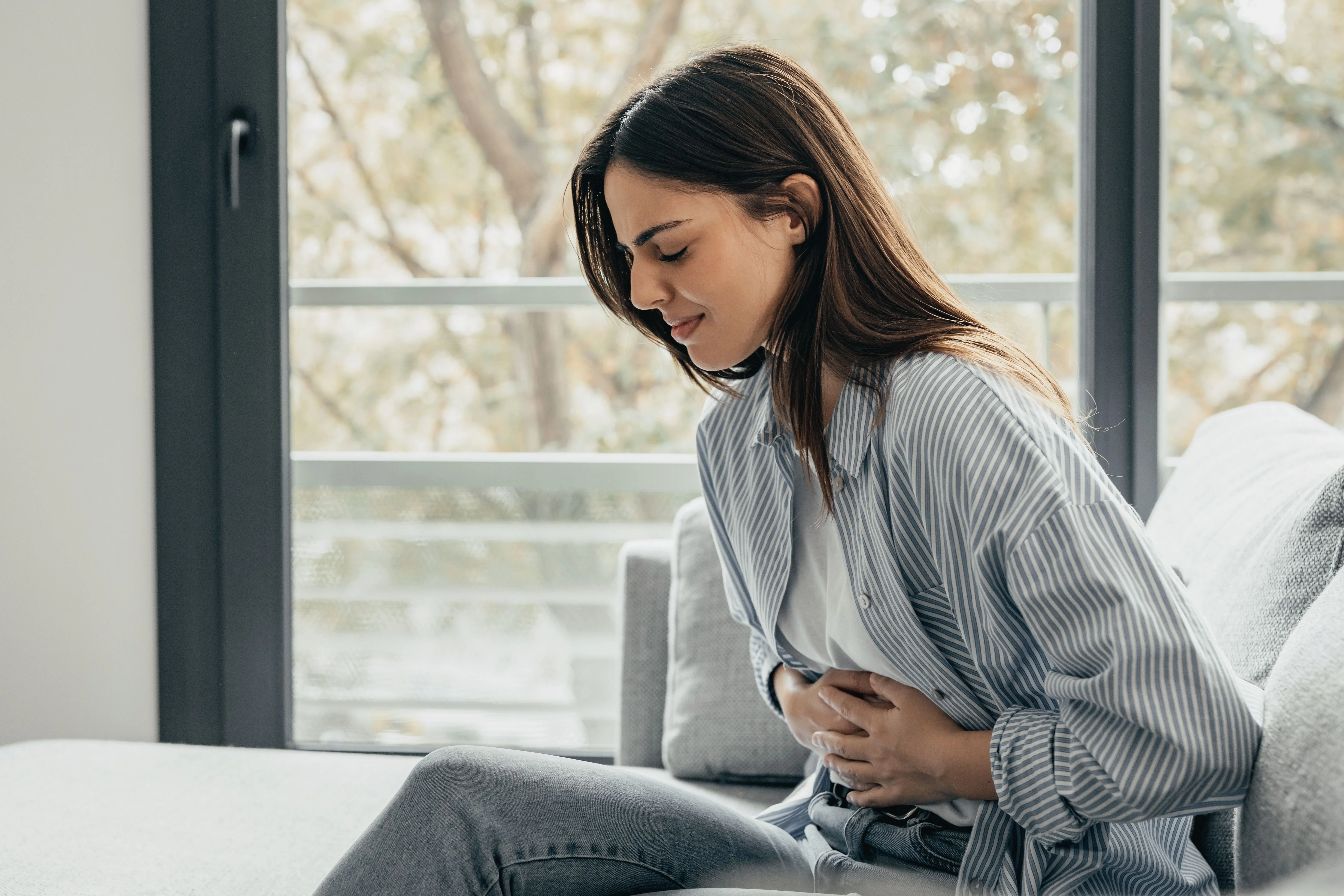 Una donna in camicia a righe è seduta su un divano, si tiene la pancia con un'espressione sofferente, luce naturale dalla finestra